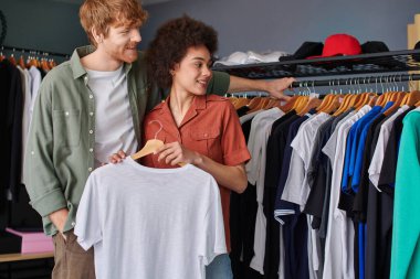 Cheerful young redhead craftsman looking at clothes on hangers while standing near african american colleague in print studio, young small business owners concept  clipart