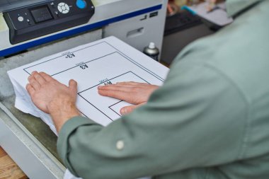 Baskı stüdyosunda işaretleme ve ekran yazdırma makinesi olan tişörtle çalışan genç zanaatkarın kırpılmış görüntüsü, müşteri odaklı küçük işletme konsepti, grafik tasarımı 