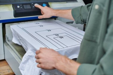 Yazılı stüdyo ve müşteri odaklı küçük işletme konseptinde işaretli tişört yanında ekran baskı makinesi kullanan bulanık genç zanaatkârın görüntüsü