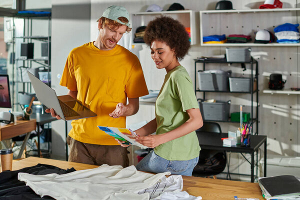 Smiling young african american craftswoman holding cloth swatches while working with colleague near laptop and clothes on table in blurred print studio, small business success concept