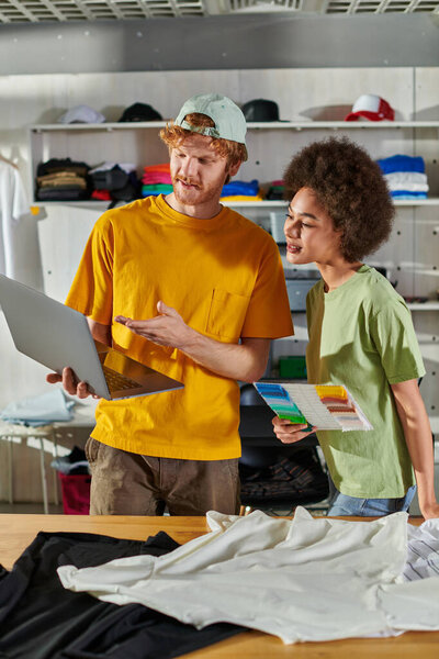 Young craftsman pointing at laptop while working with smiling african american colleague holding cloth swatches and standing near clothes in printing studio, small business success concept