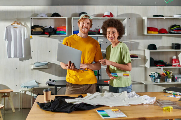 Positive young multiethnic craftspeople looking at camera while holding cloth swatches and laptop near clothes on table in print studio at background, small business success concept