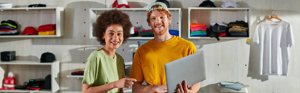 Positive multiethnic craftspeople looking at camera while working with laptop and standing near blurred clothes in print studio at background, small business success concept, banner 