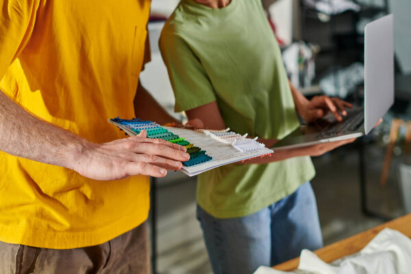 Cropped view of young craftsman holding cloth swatches while working with blurred african american colleague using laptop and standing in print studio, small business concept