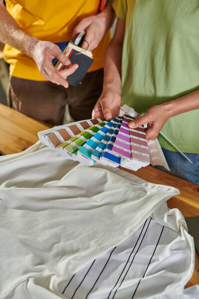 Cropped view of young african american craftswoman holding color swatches near colleague pointing with finger and standing near clothes in print studio, small business success concept