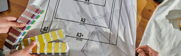 Top view of young multiethnic designers holding color swatches and printing layers while working with clothes on table in print studio, thriving small enterprise concept, banner 