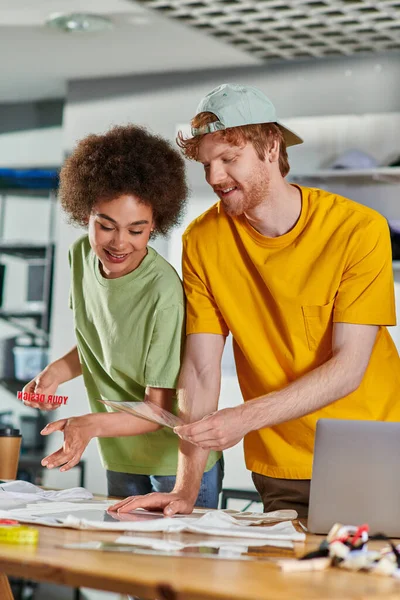 stock image Smiling craftsman holding printing layer near african american colleague, clothes and laptop on table while working in blurred print studio at background, start-up innovation concept