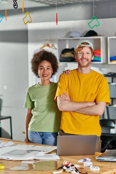 stock image Smiling young multiethnic designers looking at camera and posing while standing near laptop and clothes during project in blurred print studio at background, start-up innovation concept