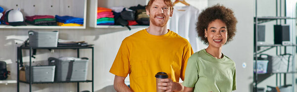 Smiling young multiethnic craftspeople with coffee to go looking at camera while standing and working on project in blurred print studio at background, start-up innovation concept, banner 