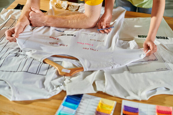 Обрезанный вид молодого африканского американского дизайнера, держащего печатный слой рядом с коллегой и футболку с надписью на столе в типографии, концепция возможности самозанятости