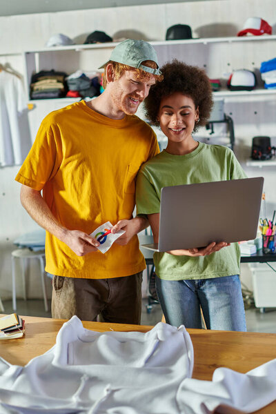 Smiling young african american designer using laptop near colleague with printing layer and clothes on table in blurred print studio at background, self-employment opportunity concept