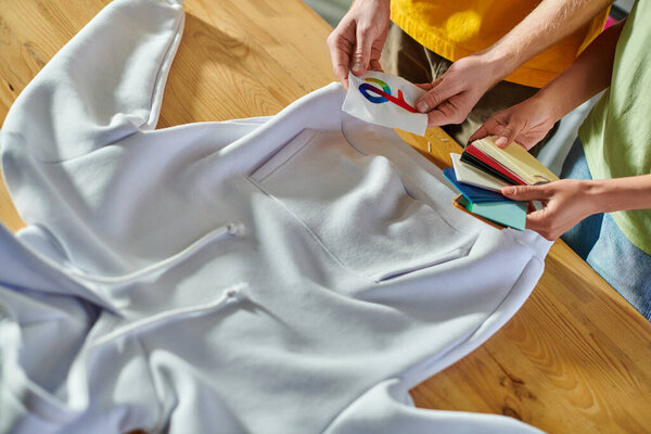 Top view of multiethnic designers holding printing layer and color swatches near clothes on table while working in print studio, self-employment opportunity concept