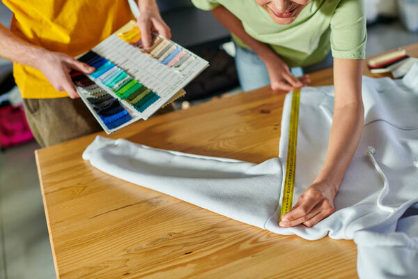 Cropped view of young african american craftswoman measuring clothes while working with colleague holding cloth samples in print studio, self-employment opportunity concept