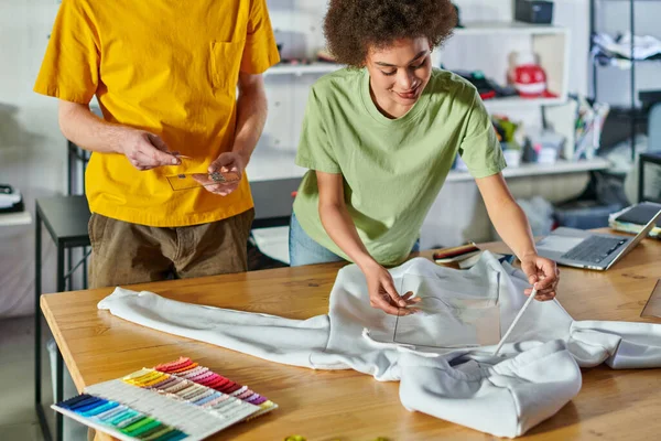 stock image Smiling young african american craftswoman holding printing layer near clothes, cloth samples and colleague while working together in print studio at background, sustainable business model concept
