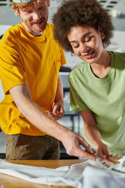 Smiling young designer holding blurred printing layer near african american colleague and clothes on table while working in blurred print studio, sustainable business model concept