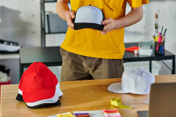 Cropped view of young craftsman holding snapback while working near tape measure and blurred laptop on table in print studio at background, small business resilience concept