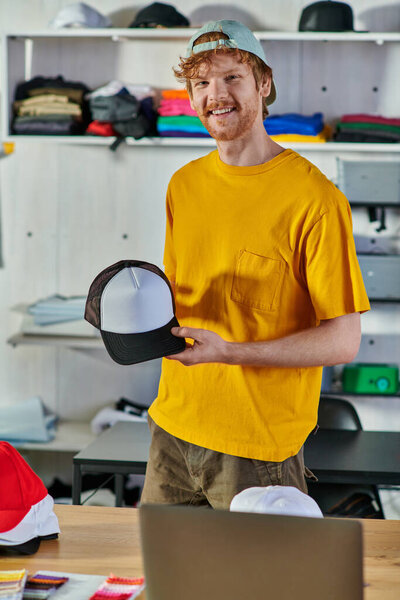 Smiling young redhead designer holding snapback and looking at camera while working near laptop and cloth samples in blurred print studio, small business resilience concept