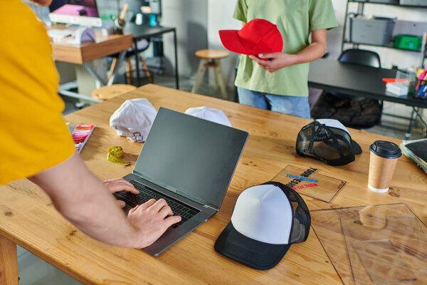 Cropped view of young craftsman using laptop near coffee to go and african american colleague with snapback in print studio at background, small business resilience concept