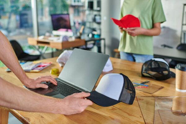 Cropped view of young designer using laptop and holding snapback and working near blurred african american colleague at background in print studio, small business resilience concept