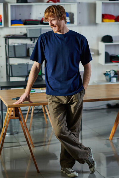 Smiling young redhead craftsman in casual clothes holding hand in pocket while standing near working table in print studio, hands-on entrepreneurship concept 