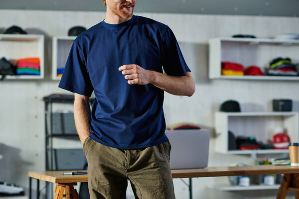 Обрезанный вид улыбающегося молодого ремесленника в повседневной одежде, держащегося за карман, стоя рядом с рабочим столом в типографии, самодельная концепция успеха 