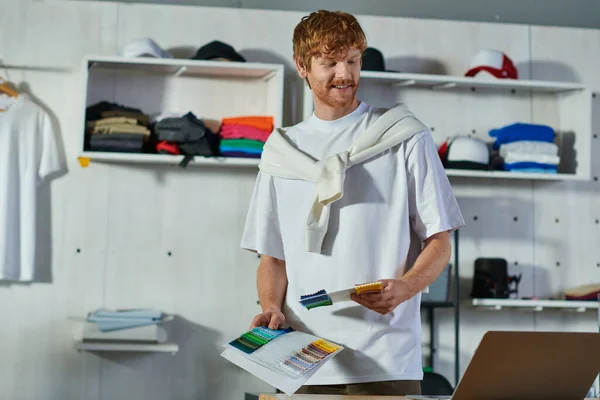 Smiling Young Redhead Craftsman Holding Cloth Samples While Looking Laptop — Stock Photo, Image