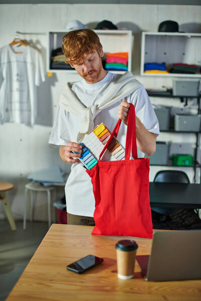 Молодой рыжий ремесленник в повседневной одежде, держа образцы ткани рядом с плечевой сумкой и приборы на столе в размытой типографии на заднем плане, самодельная концепция успеха 
