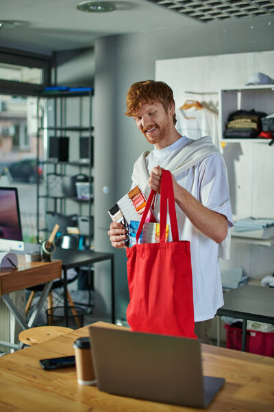 Веселый молодой рыжий ремесленник в повседневной одежде, держа образцы ткани и плечевую сумку, работая рядом с устройствами и кофе, чтобы пойти в типографию, самодельная концепция успеха