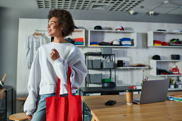 Smiling young african american craftswoman holding shoulder bag and looking away while standing near devices and coffee to go in print studio, enthusiastic business owner working in workshop