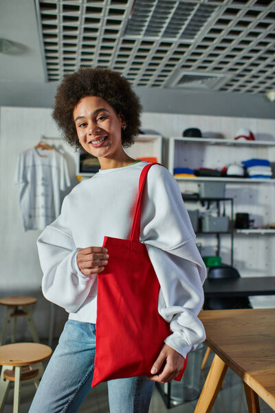 Cheerful young african american craftswoman holding shoulder bag and looking at camera while standing in blurred print studio, enthusiastic business owner working in workshop
