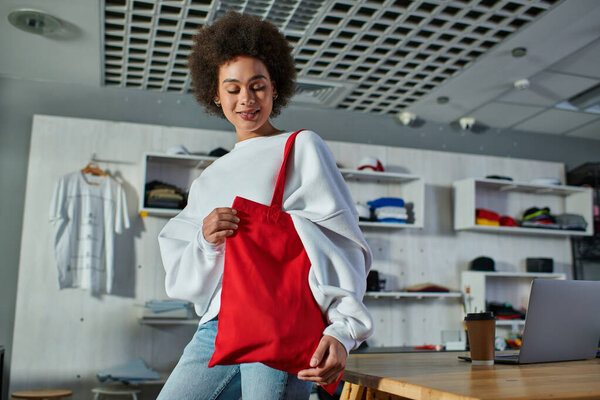 Joyful young african american craftswoman looking at shoulder bag while standing near coffee to go and laptop on table in print studio, enthusiastic business owner working in workshop