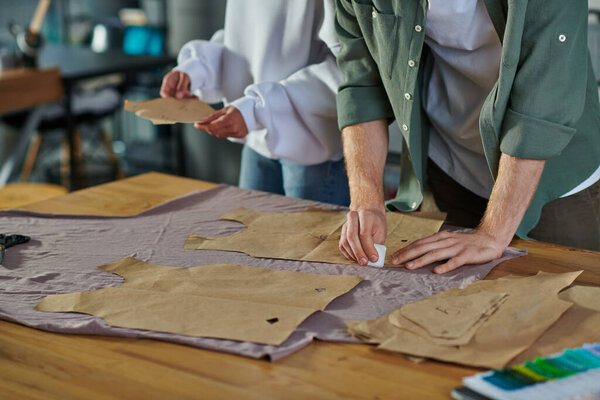 Cropped view of craftsman marking cloth near sewing patterns and blurred african american colleague while working in print studio, collaborative business owners working together 