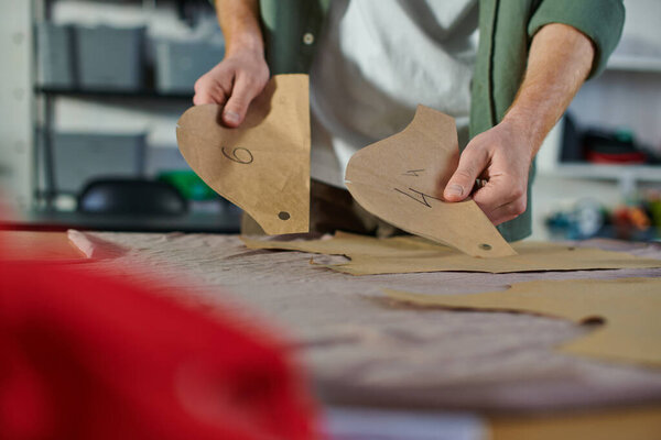 Cropped view of young craftsman holding sewing patterns near blurred cloth on table in print studio at background, multitasking business owner managing multiple project