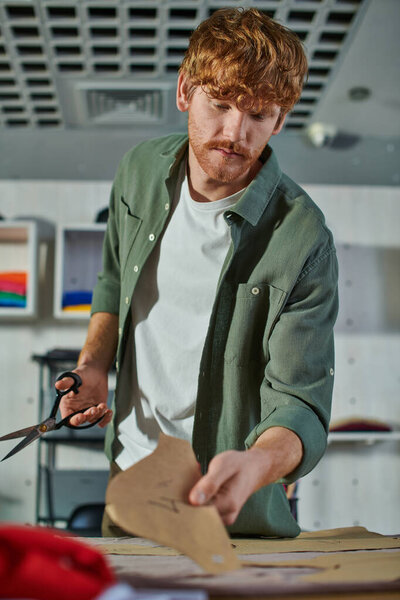 Young redhead craftsman holding scissors and blurred sewing pattern near fabric on table in blurred print studio at background, multitasking business owner managing multiple project