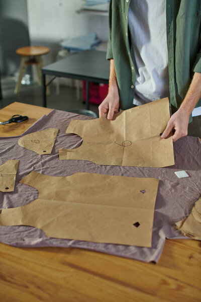 Cropped view of young craftsman putting sewing pattern on fabric near chalk and scissors on table in print studio, multitasking business owner managing multiple project