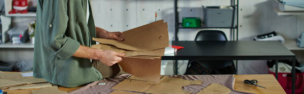 Cropped view of young craftsman holding sewing patterns near cloth and scissors on table in blurred print studio, multitasking business owner managing multiple project, banner 
