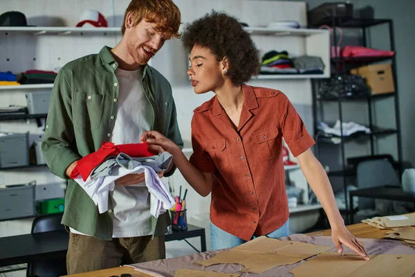 stock image Young african american craftswoman looking at clothes in hands of cheerful colleague and working with fabric and sewing patterns in print studio, ambitious young entrepreneurs concept 