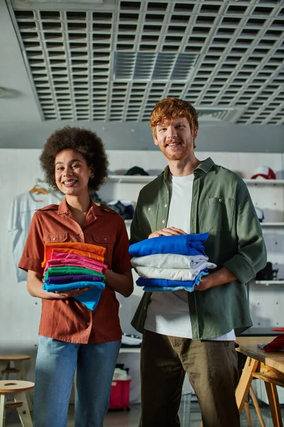 stock image Cheerful young interracial craftspeople holding clothes and looking at camera while standing in blurred print studio at background, ambitious young entrepreneurs concept 