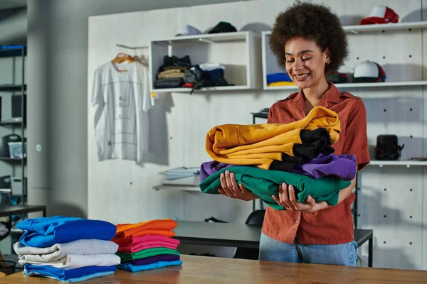 Positive young african amercan craftswoman holding clothes near working table while standing in blurred print studio at background, customer-focused entrepreneur concept