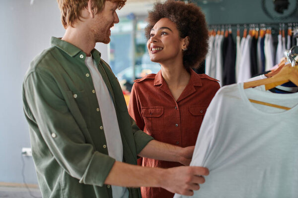 Positive young african american craftswoman looking at redhead colleague while standing near clothes on hangers in blurred print studio, young small business owners concept 