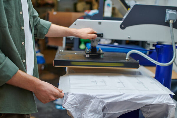 Cropped view of craftsman holding layer on t-shirt and working with screen printing machine in blurred workshop, customer-focused small business concept