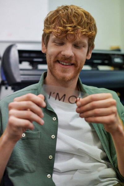 Smiling young redhead craftsman holding layer with I'm ok lettering while working near screen printing machine in workshop, small business owner working on project 