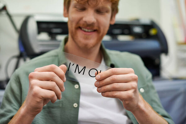 Blurred young craftsman smiling and holding layer with I'm ok lettering near screen printing machine in workshop, small business owner working on project 
