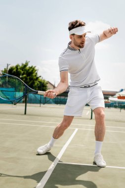 sporty man in sports visor holding racket and playing tennis on court, training and motivation clipart