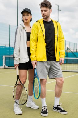 spor, şık bir çift tenis kortunda raketlerle duruyorlar, kadın ve erkek şık kıyafetler giyiyorlar.