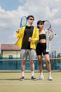 Spor motivasyonu ve moda, güneş gözlüklü kadın ve erkek tenis kortunda raket ve top tutuyorlar.