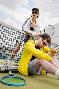 Tenis ağının yanında poz veren güneş gözlüklü, raket ve top tutan aktif giyimli bir çift.