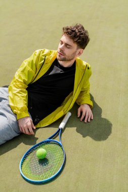 Üstten atış, kortta erkek tenisçi, tenis topu ve raketin yanında dinleniyor, aktif giysi