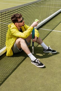 Güneş gözlüklü yakışıklı adam tenis ağının yanında oturuyor, elinde top ve raket, erkek tenisçi.