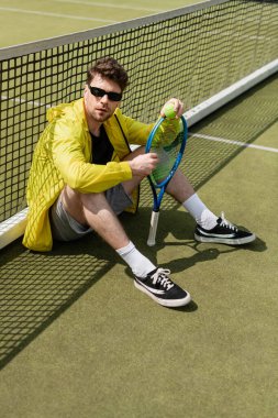 Güneş gözlüklü erkek tenisçi tenis ağının yanında oturuyor, top ve raket tutuyor, spor ve tarz sahibi.